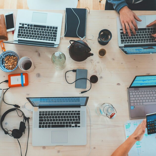 Arbeitsplatz mit mehreren Laptops, Notizbuch, Smartphone, Kopfh&ouml;rern, Getr&auml;nken und Snacks; mehrere Personen arbeiten gemeinsam an einem Tisch.