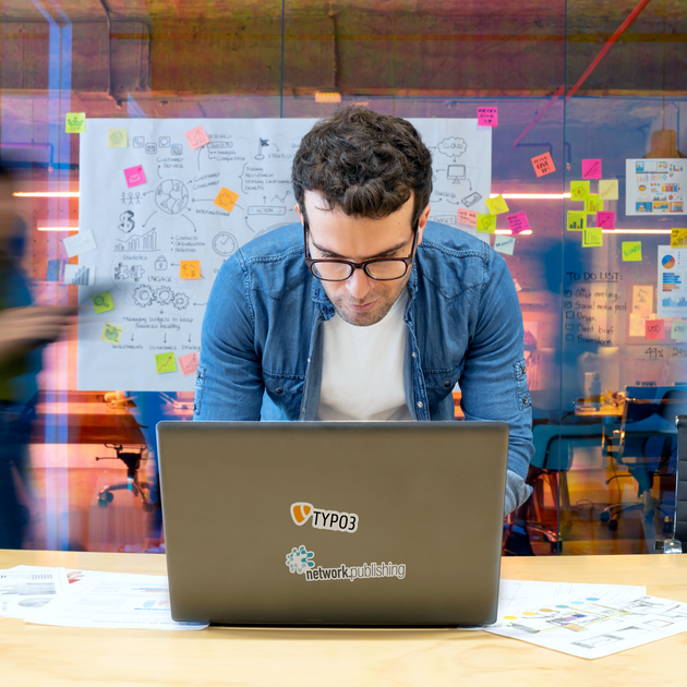 Ein Mann arbeitet an einem Laptop in einem belebten Büro, mit einer Tafel und Postits im Hintergrund.